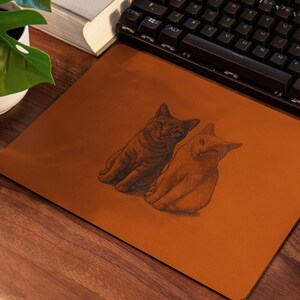 Pode incluir: Um mousepad retangular castanho com uma ilustra&ccedil;&atilde;o detalhada de dois gatos. Os gatos s&atilde;o retratados sentados com os olhos fechados. O mousepad est&aacute; em uma mesa de madeira ao lado de um teclado e um mouse.