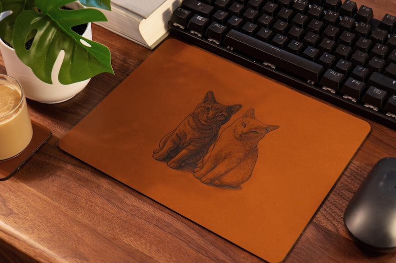 Pode incluir: Um mousepad retangular castanho com uma ilustra&ccedil;&atilde;o detalhada de dois gatos. Os gatos s&atilde;o retratados sentados com os olhos fechados. O mousepad est&aacute; em uma mesa de madeira ao lado de um teclado e um mouse.