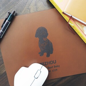 Pode incluir: Um mousepad de couro castanho com uma ilustra&ccedil;&atilde;o de c&atilde;o preto e o texto "MIZHOU Best Baby 1229". Um rato de computador branco e uma caneta tamb&eacute;m s&atilde;o vis&iacute;veis.
