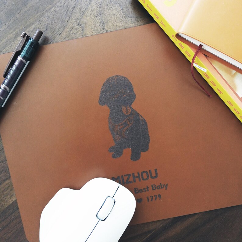 Pode incluir: Um mousepad de couro castanho com uma ilustra&ccedil;&atilde;o de c&atilde;o preto e o texto "MIZHOU Best Baby 1229". Um rato de computador branco e uma caneta tamb&eacute;m s&atilde;o vis&iacute;veis.