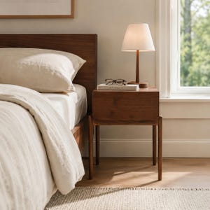 Puede incluir: Una mesita de noche de madera oscura con cuatro patas delgadas y un caj&oacute;n. Encima hay una l&aacute;mpara de mesa con pantalla c&oacute;nica blanca y base de madera oscura, un libro blanco titulado Kinfolk y unas gafas de montura negra sobre el libro.