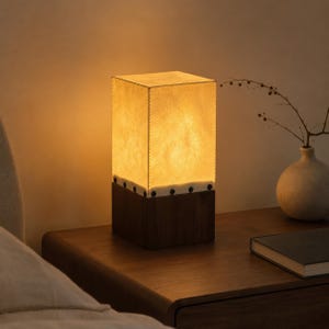 May include: A rectangular table lamp featuring a warm, glowing, textured off-white shade with visible stitching along the edges. The shade is secured to a solid, dark brown wooden base by a series of small, evenly spaced dark metal rivets.