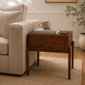Puede incluir: Una mesa auxiliar rectangular de madera oscura con cuatro patas delgadas. Sobre la superficie plana hay una taza de cer&aacute;mica beige, un libro titulado The Kinfolk Home y un tel&eacute;fono inteligente negro. La mesa est&aacute; junto a un sof&aacute; beige.