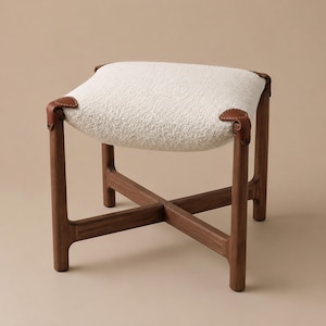 May include: A stool featuring a white textured fabric seat cushion secured by brown leather straps to a dark wood frame. The base consists of four legs connected by a cross-shaped support structure, providing a stable foundation for the seat.