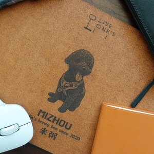 Pode incluir: Um caderno castanho com uma ilustra&ccedil;&atilde;o de c&atilde;o preto e o texto "MIZHOU". Um rato de computador branco, uma carteira preta e um caderno laranja tamb&eacute;m s&atilde;o vis&iacute;veis. Uma caneta est&aacute; no canto superior esquerdo.