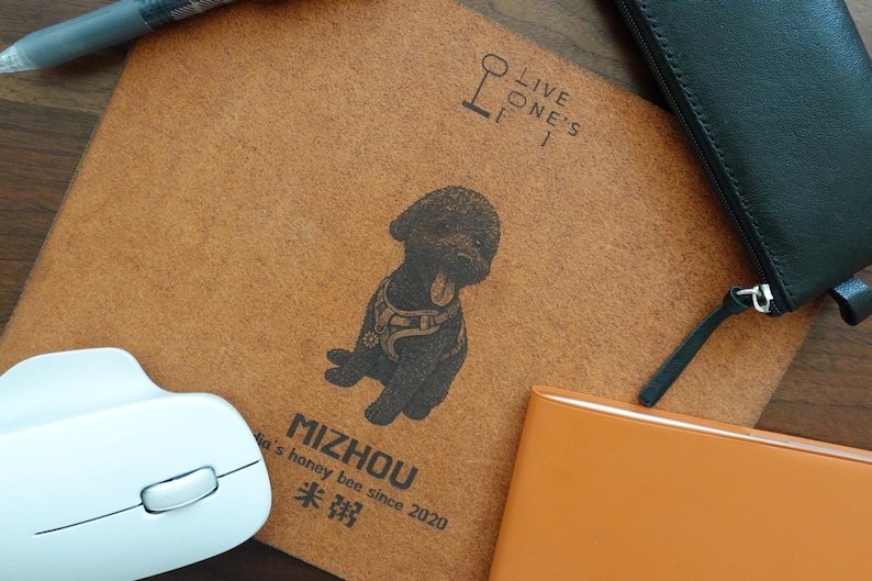 Pode incluir: Um caderno castanho com uma ilustra&ccedil;&atilde;o de c&atilde;o preto e o texto "MIZHOU". Um rato de computador branco, uma carteira preta e um caderno laranja tamb&eacute;m s&atilde;o vis&iacute;veis. Uma caneta est&aacute; no canto superior esquerdo.