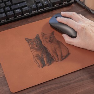 Pode incluir: Um mousepad retangular marrom com duas ilustra&ccedil;&otilde;es de gatos. Os gatos s&atilde;o desenhados em um estilo detalhado e sombreado. Um mouse sem fio preto est&aacute; no mousepad e um teclado &eacute; vis&iacute;vel ao fundo.