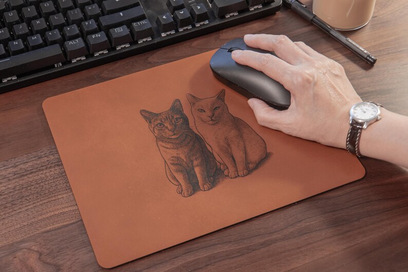 Pode incluir: Um mousepad retangular marrom com duas ilustra&ccedil;&otilde;es de gatos. Os gatos s&atilde;o desenhados em um estilo detalhado e sombreado. Um mouse sem fio preto est&aacute; no mousepad e um teclado &eacute; vis&iacute;vel ao fundo.