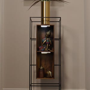 May include: A tall, black metal etagere with a gold vase holding a plant. The shelves display a Buddha statue and ceramic vases. The etagere is a decorative piece for home decor.
