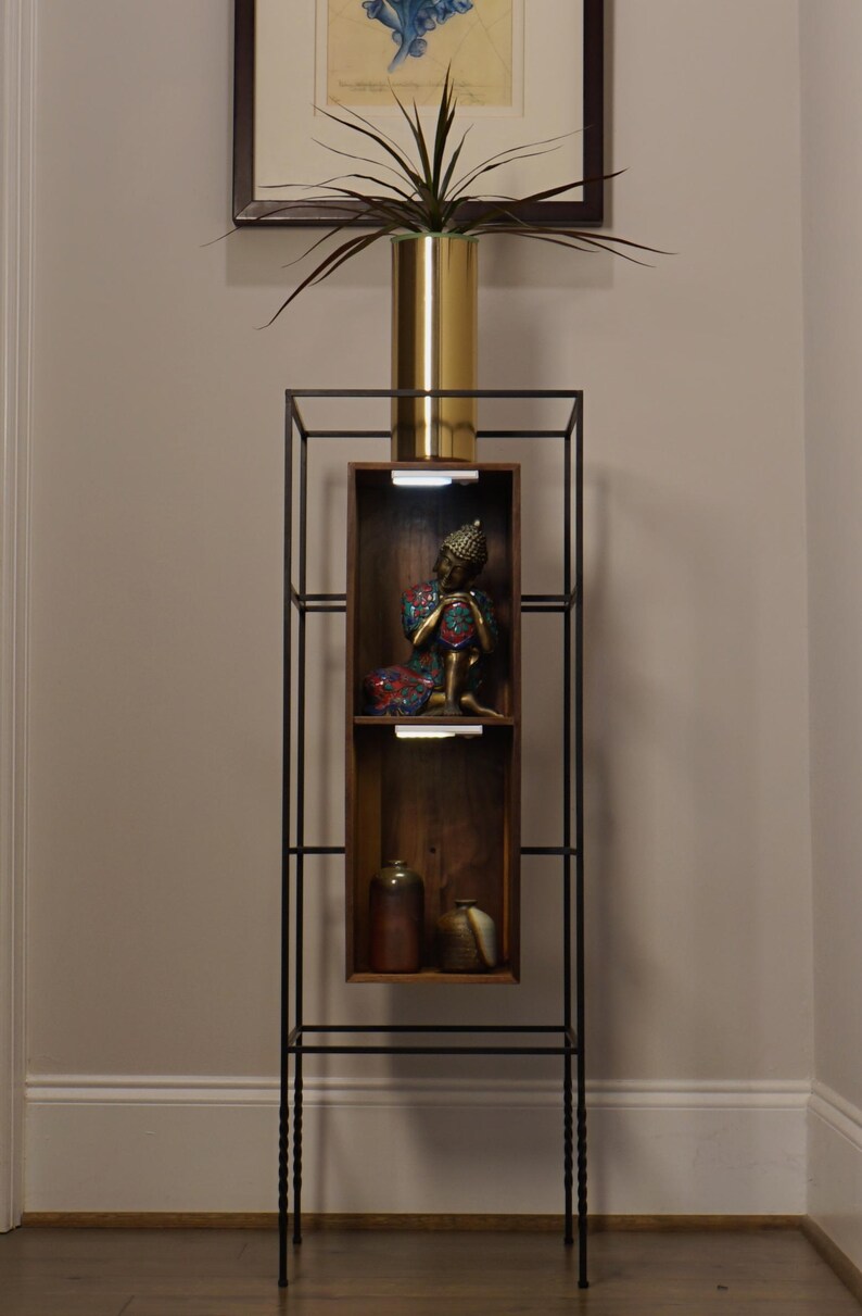 May include: A tall, black metal etagere with a gold vase holding a plant. The shelves display a Buddha statue and ceramic vases. The etagere is a decorative piece for home decor.
