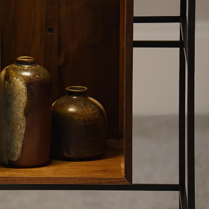 May include: Two ceramic vases with a brown and gold glaze sit on a wooden shelf. The vases have a rounded shape and a narrow opening. The shelf is part of a dark wooden cabinet with a black metal frame.