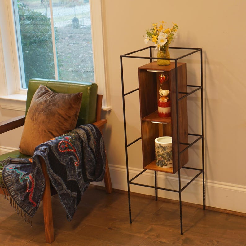 May include: A tall, black metal and wood shelf unit with three shelves. The shelves hold a vase of yellow and white flowers, a teddy bear figurine, and a decorative ceramic container. The unit is in a room with a green chair and a window.