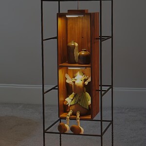May include: A decorative display shelf with a dark metal frame and wooden shelves. The shelves contain a stuffed moose toy, ceramic vases, and a small bird figurine. The interior of the shelves are illuminated.