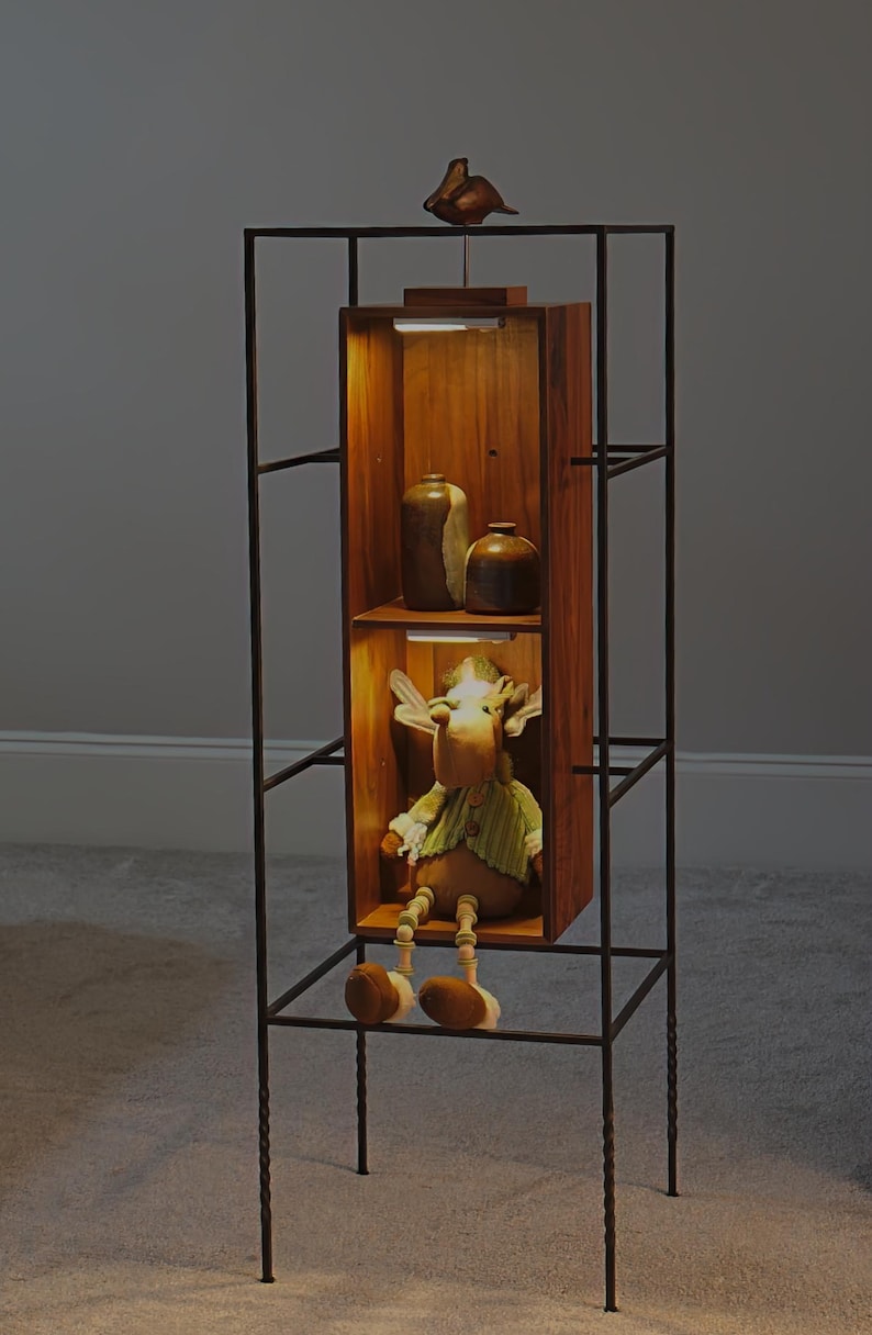 May include: A decorative display shelf with a dark metal frame and wooden shelves. The shelves contain a stuffed moose toy, ceramic vases, and a small bird figurine. The interior of the shelves are illuminated.