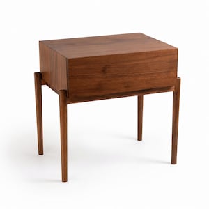 Puede incluir: Una mesita de noche rectangular de madera con una veta marr&oacute;n intensa, una superficie superior plana y un compartimento de caj&oacute;n frontal. La pieza se apoya sobre cuatro patas cil&iacute;ndricas y delgadas que se extienden verticalmente desde la base.