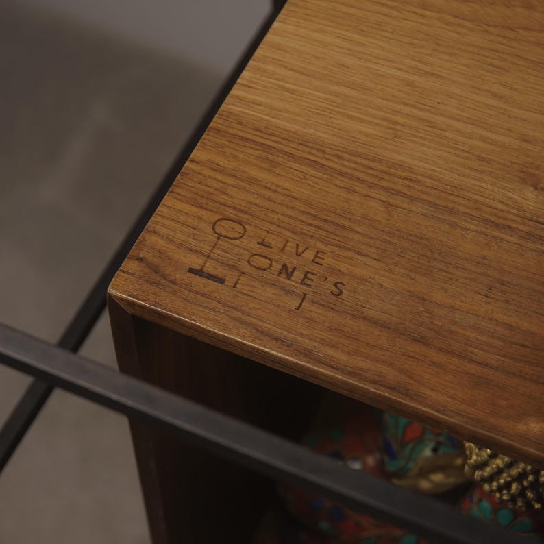 May include: Close-up of a wooden table corner with the text "LIVE ONE'S" etched into the surface. The wood has a warm, brown tone with visible grain. A dark metal frame is partially visible, suggesting a larger furniture piece.