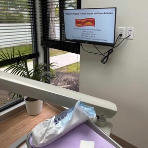 May include: A dental surgery scene featuring a monitor showing the text "Plaque is Plaque: In Your Mouth and Your Arteries." The screen displays an image of plaque. A dental chair and tray are visible in the foreground.