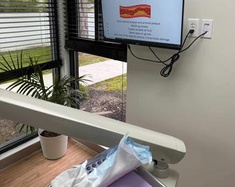 May include: A dental office setting with a monitor displaying the text "Plaque is Plaque: In Your Mouth and Your Arteries." The monitor shows an illustration of plaque buildup. A dental chair and tray are in the foreground.