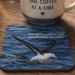 Könnte beinhalten: Ein Keramikbecher mit gesprenkelter Oberfläche und blauem Rand, der den Text "SURVIVING WORK ONE COFFEE AT A TIME" mit einem Herzsymbol zeigt. Daneben zeigt ein quadratischer Untersetzer einen Albatros über Wasser mit dem Text "Current status: running on caffeine and questionable decisions."