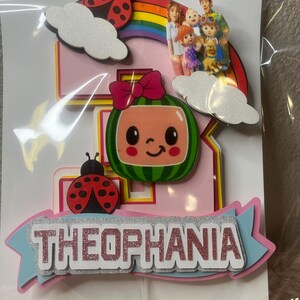 May include: A pink and blue birthday cake topper with the name "THEOPHANIA" in glittery letters. The topper features a watermelon character, ladybugs, clouds, a rainbow, and a family illustration.