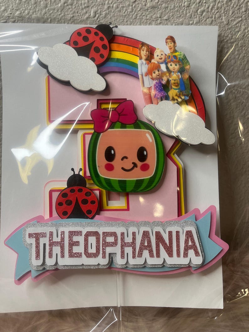 May include: A pink and blue birthday cake topper with the name "THEOPHANIA" in glittery letters. The topper features a watermelon character, ladybugs, clouds, a rainbow, and a family illustration.