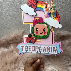 May include: A pink and yellow number five cake topper with a watermelon character, ladybugs, and a rainbow. The topper has the name "THEOPHANIA" in silver glitter. A family of characters is on top of the rainbow.