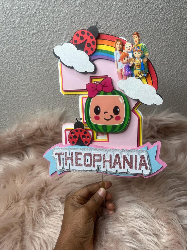 May include: A pink and yellow number five cake topper with a watermelon character, ladybugs, and a rainbow. The topper has the name "THEOPHANIA" in silver glitter. A family of characters is on top of the rainbow.