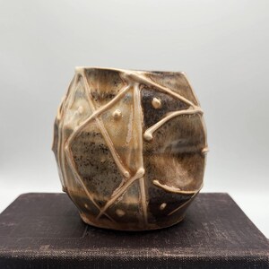 May include: A round ceramic vase with a mottled brown and tan glaze, featuring a raised, cream-colored geometric lattice pattern across its surface. The vessel has a wide, open top and a tapered base, showcasing a textured, hand-crafted appearance.