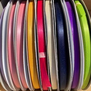 May include: A collection of colorful satin ribbons in various shades, including pink, yellow, red, navy, purple, and green. The ribbons are neatly arranged on cardboard spools, offering a variety of options for crafting and gift wrapping.