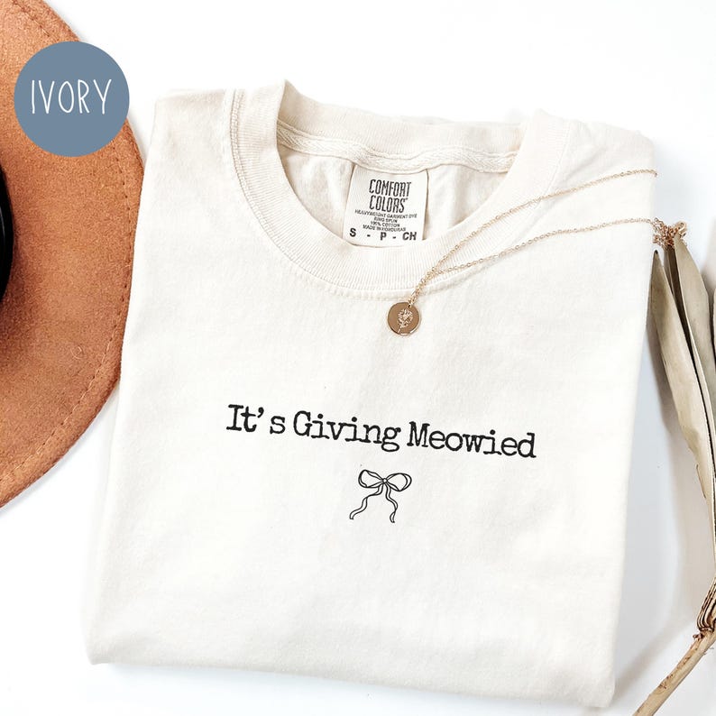 May include: Ivory-colored t-shirt with the phrase "It's Giving Meowied" in black script, accompanied by a bow illustration. A gold necklace with a pendant rests on the shirt. The shirt is folded, and a brown hat is partially visible.