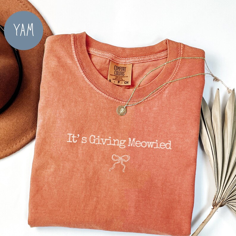 May include: A coral-colored Comfort Colors t-shirt with the phrase "It's Giving Meowied" in white script and a bow graphic. A gold necklace with a pendant rests on the shirt. A brown hat and a dried palm leaf are in the background.