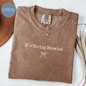 May include: Espresso-colored t-shirt with the phrase "It's Giving Meowied" and a bow graphic. The shirt has a crew neck and a Comfort Colors tag. A silver necklace with a pendant is draped across the shirt.