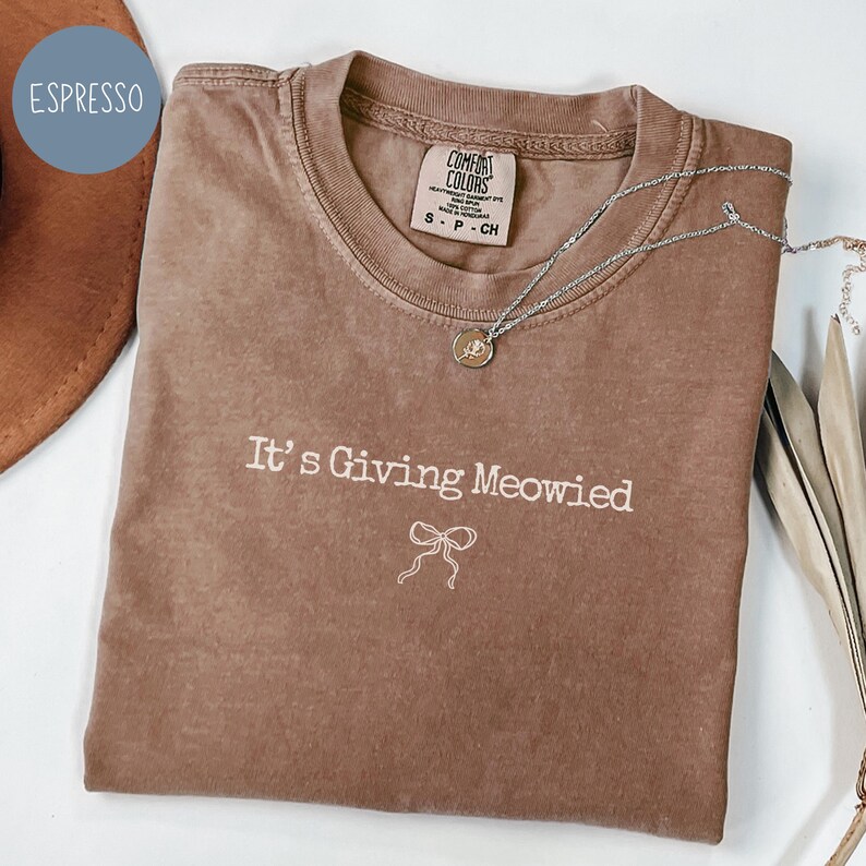May include: Espresso-colored t-shirt with the phrase "It's Giving Meowied" and a bow graphic. The shirt has a crew neck and a Comfort Colors tag. A silver necklace with a pendant is draped across the shirt.