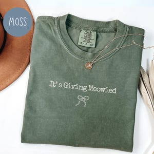 May include: A moss green Comfort Colors t-shirt with the phrase "It's Giving Meowied" and a bow graphic. A gold-colored necklace with a round pendant rests on the shirt. The shirt is folded and displayed with a brown hat.
