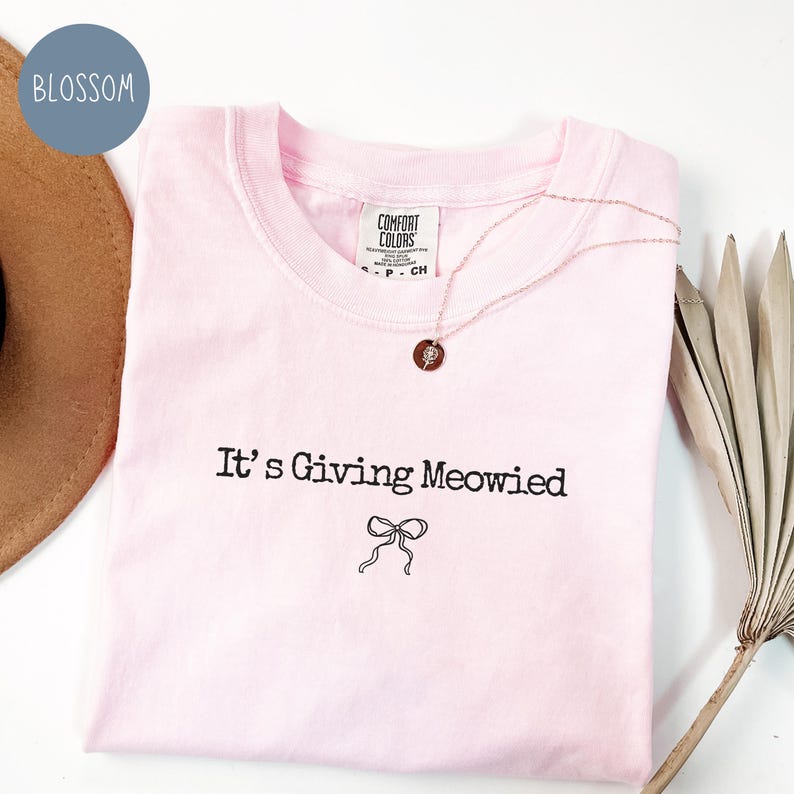 May include: A light pink t-shirt with the phrase "It's Giving Meowied" in black script, along with a bow graphic. A rose gold necklace with a pendant rests on the shirt. The shirt has a Comfort Colors tag.