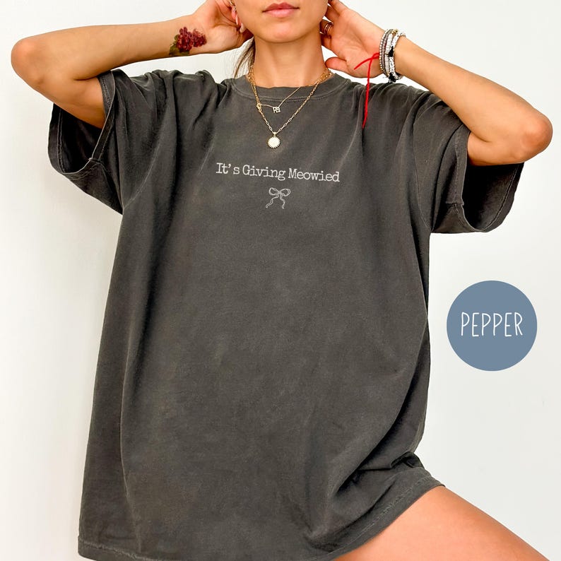 May include: Dark gray oversized t-shirt with the phrase "It's Giving Meowied" embroidered in white, along with a bow graphic. The shirt has short sleeves and a relaxed fit. A blue circle with the word "PEPPER" is in the lower right corner.