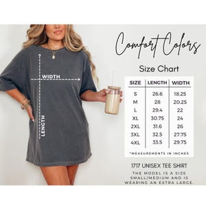 May include: A dark gray unisex tee shirt with a size chart. The size chart shows the length and width measurements in inches for sizes S to 4XL. The text on the shirt reads "1717 Unisex Tee Shirt". The model is wearing an extra large.