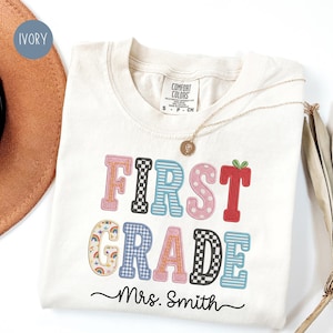 May include: An ivory t-shirt featuring the text FIRST GRADE in colorful, patterned block letters, including checkered, striped, polka dot, and rainbow prints. Below the text, the name Mrs. Smith is printed in a black cursive script font.