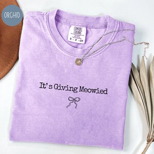 May include: A light purple t-shirt with the phrase "It's Giving Meowied" and a bow graphic. A silver necklace with a round pendant rests on the shirt. The shirt is labeled "Comfort Colors" and is size small.