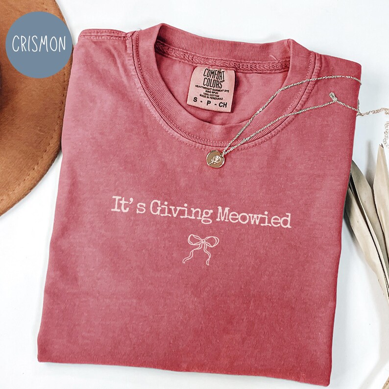 May include: A crimson-colored Comfort Colors t-shirt with the phrase "It's Giving Meowied" in white script, along with a bow graphic. A silver necklace with a pendant rests on the shirt. The shirt is size small.