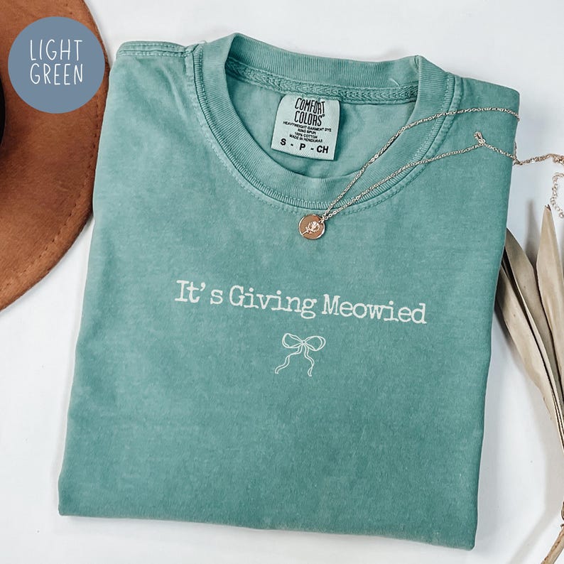 May include: A light green t-shirt with the phrase "It's Giving Meowied" in white script and a bow graphic. A silver necklace with a pendant is draped across the shirt. The shirt is folded and displayed with a brown hat and dried leaves.