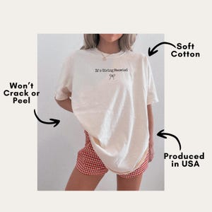 May include: Cream-colored t-shirt with the phrase "It's Giving Meowied" and a bow graphic. The shirt is made of soft cotton and produced in the USA. The design is made to not crack or peel.
