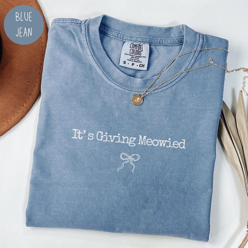 May include: A blue jean Comfort Colors t-shirt with the phrase "It's Giving Meowied" embroidered in white, along with a bow graphic. A gold necklace with a pendant rests on the shirt.