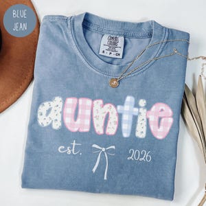 May include: Blue jean-colored t-shirt with the word "auntie" in a floral and gingham design. The shirt also features the text "est. 2026" and a bow graphic. A gold necklace with a pendant is draped across the shirt.