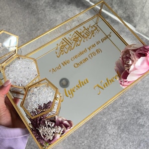 May include: A mirrored tray with a gold frame, featuring gold Arabic calligraphy and the English text "And We created you in pairs" from the Quran. The names "Ayesha" and "Nabee" are also inscribed. Decorative flowers and hexagonal glass containers filled with crystals are included.