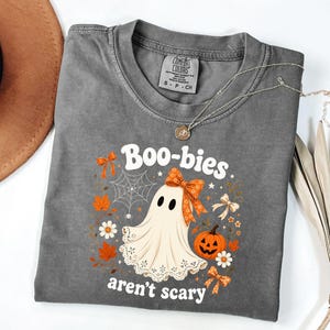May include: A charcoal gray t-shirt featuring a graphic of a ghost wearing an orange patterned bow, accompanied by a jack-o'-lantern, autumn leaves, a spider web, and small flowers. The text reads "Boo-bies aren't scary" in white lettering.