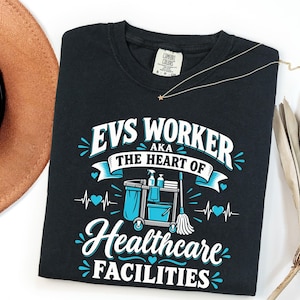 May include: A black t-shirt featuring white and light blue graphic text that reads EVS WORKER AKA THE HEART OF HEALTHCARE FACILITIES. The design includes a cleaning cart, a mop, and heart symbols integrated into a heartbeat line graphic.