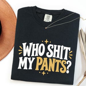 May include: A black short-sleeved t-shirt featuring the white and gold text "WHO SHIT MY PANTS?" printed on the front. The design includes small gold star and sunburst accents above and below the text. The shirt has a Comfort Colors brand label.
