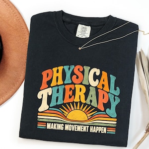 May include: A black t-shirt featuring the text PHYSICAL THERAPY in a multi-colored, retro-style font. Below the text is a graphic of a rising sun with horizontal stripes. The text MAKING MOVEMENT HAPPEN is printed in white at the bottom.