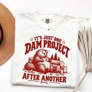 May include: A white t-shirt featuring a red graphic of a beaver sitting on logs, holding a mug with a tree icon. The text reads, "IT'S JUST ONE DAM PROJECT AFTER ANOTHER." The shirt has a Comfort Colors brand label at the inner neckline.